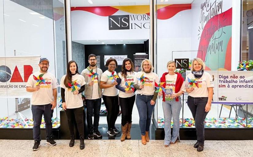 Seis mulheres e dois homens posam para a foto. Todos estão estão de pé, em frente a uma vitrine, e seguram cataventos coloridos. Uma faixa ao lado deles apresenta o seguinte texto: Diga não ao trabalho infantil e sim à aprendizagem.