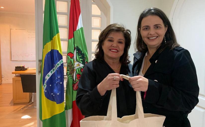 Duas mulheres em pé e sorridentes segiram uma sacola. Ao fundo, as bandeiras do Brasil e de Santa Catarina