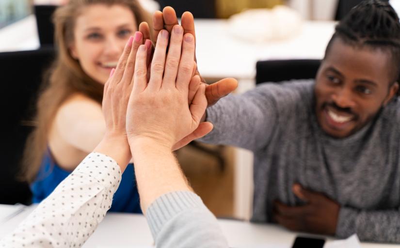 close nas mãos unidas de pessoas que estão em uma reunião. no fundo, desfocado, é possível ver o rosto de uma mulher e de um homem sorrindo