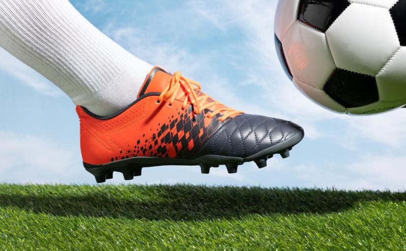 Imagem ilustrativa da visão lateral de um pé com chuteira prestes a chutar uma bola de futebol em um campo gramado