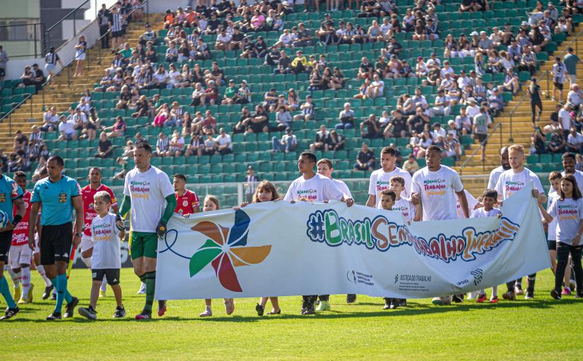 Jogadores de futebol entram em campo, ao lado de crianças, segurando uma faixa contendo o texto Brasil Sem Trabalho Infnatil