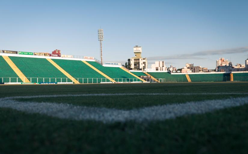 Foto de um estádio de futebol tirada do corner de escanteio, mostrando gramado e parte das arquibancadas, que têm cadeiras verdes