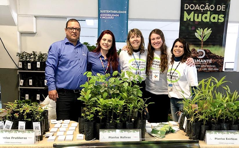 Um homem e quatro mulheres estão em pé e posando para a foto. Na frente deles uma mesa com mudas de plantas