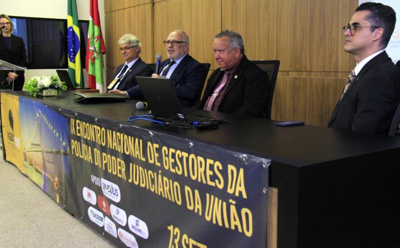 Em uma mesa retangular estão sentados quatro homens brancos, trajando terno e gravata, com um banner a frente que diz 9