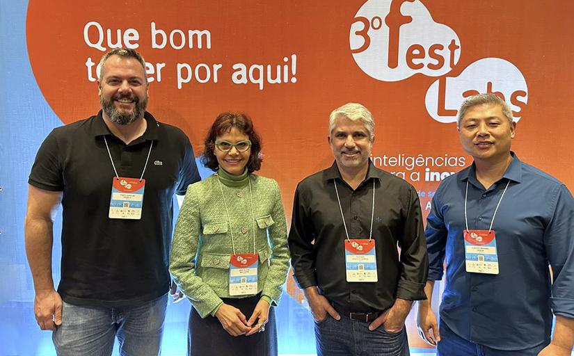 Três homens e uma mulher posam em frente a um banner de cor laranja, com os textos: 3º FestLabs, que bom te ver por aqui