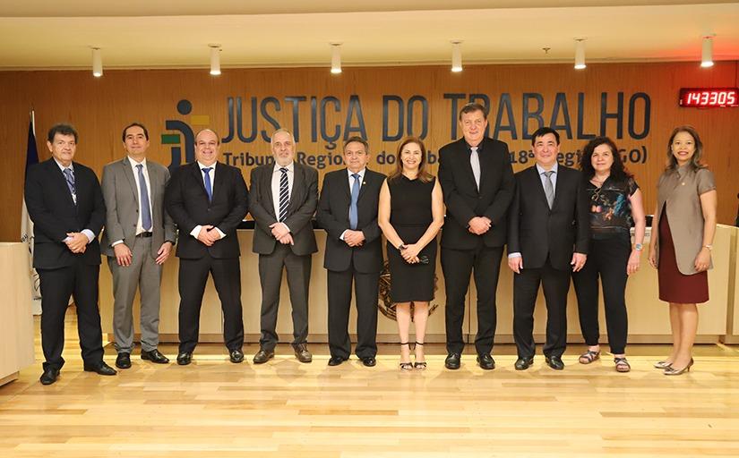 Sete homens, todos de terno, e três mulheres posam para foto lado a lado, em pé. Eles estão posicionados sob seis spots de lâmpadas amarelas. Ao fundo, se lê a expressão Justiça do Trabalho plotada em um painel de MDF