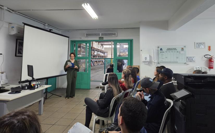 Foto mostra uma mulher morena com uma roupa escura em frente a uma lousa branca, falando diante de um grupo de pessoas