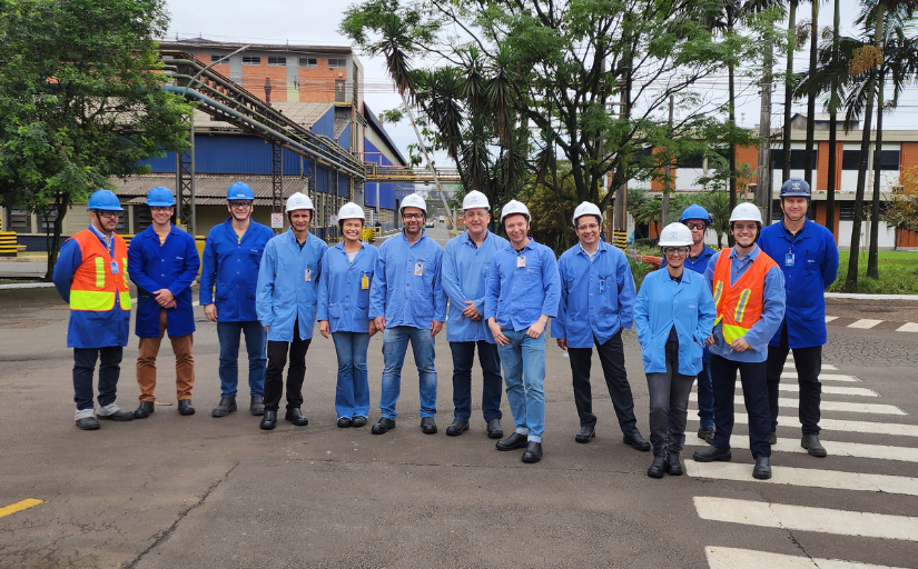 Grupo de pessoas, homens e mulheres, estão em pé posando para a foto, com roupa azul e capacete, na parte externa de uma fábrica