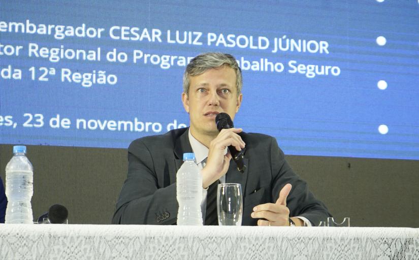 Homem de cabelos grisalhos, olhos azuis e pele branca, vestindo um terno azul, sentado em frente a uma mesa segurando um microfone