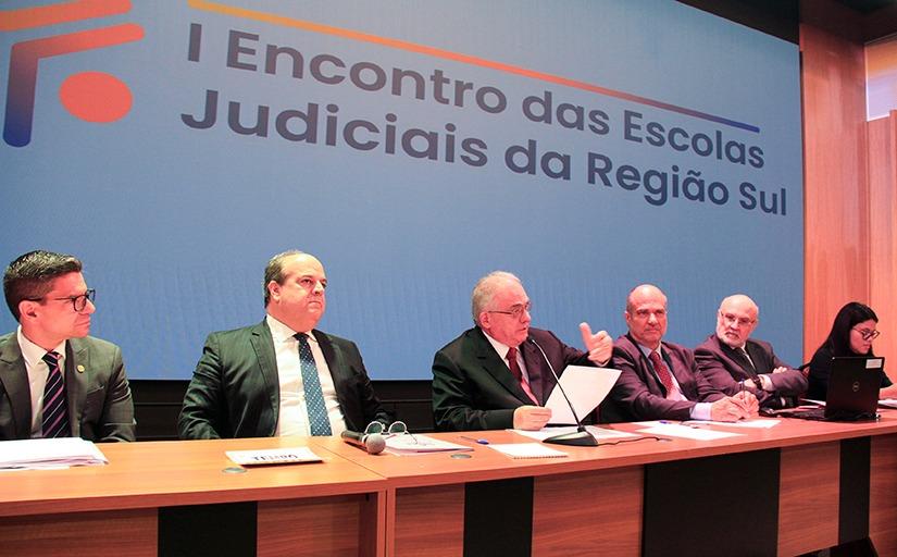 Cinco homens de terno e gravata e uma mulher de preto estão sentados em uma mesa retangular no palco de um auditório. Atrás um telão com a imagem: 1º Encontro das Escolas Judiciais da Região Sul