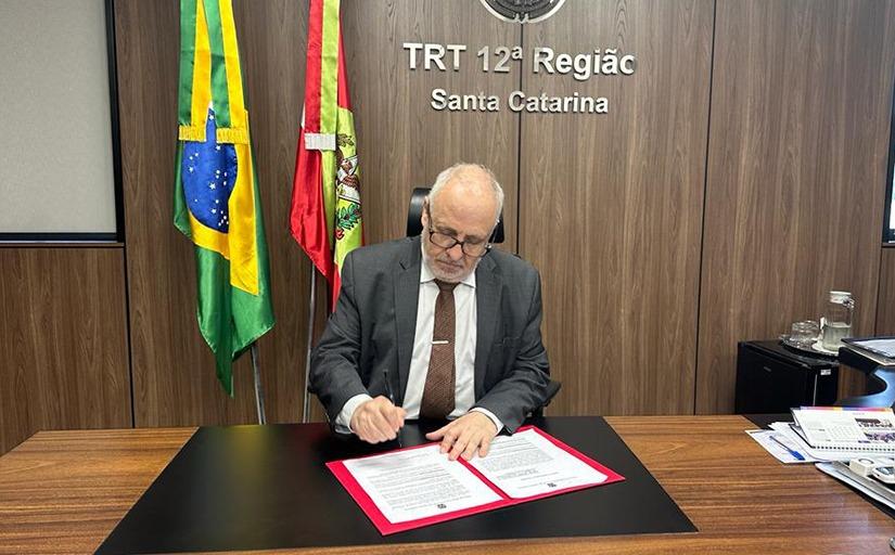 Homem de terno escuro, sentado, assina um documento. Ao fundo, um MDF marrom, as bandeiras do Brasil e de Santa Catarina e o texto, em letras de metal dourado: TRT da 12ª Região - Santa Catarina