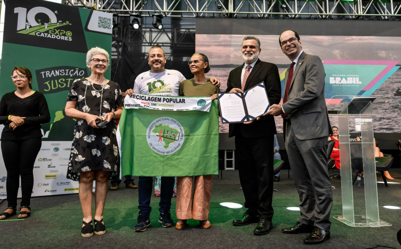 Cinco pessoas em pé, segurando uma bandeira verde com um mapa do Brasil.  