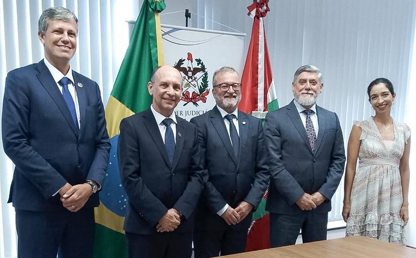 Foto posada de quatro homens de terno escuro e uma mulher de vestido claro, em pé, lado a lado. Ao fundo, as bandeiras do Brasil, de Santa Catarina e um brasão do Poder Judiciário