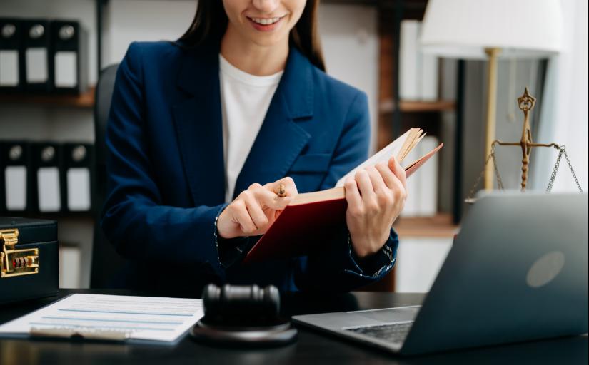 mulher de cabelos escuros vestida com terno azul e camiseta branca, sentada atrás de uma mesa com um notebook, um martelo de juiz e alguns papéis