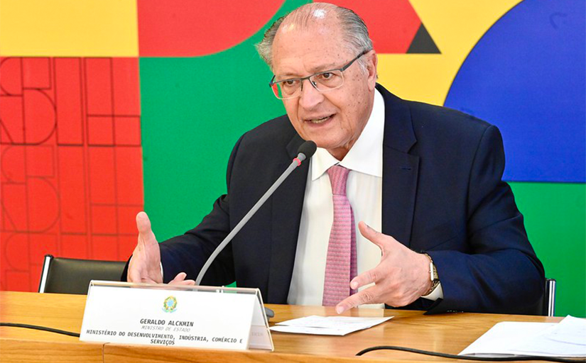 Geraldo Alckmin, um homem branco, usando óculos, trajando terno azul marinho, camisa branca e gravata rosa, está sentado em uma mesa falando em um microfone