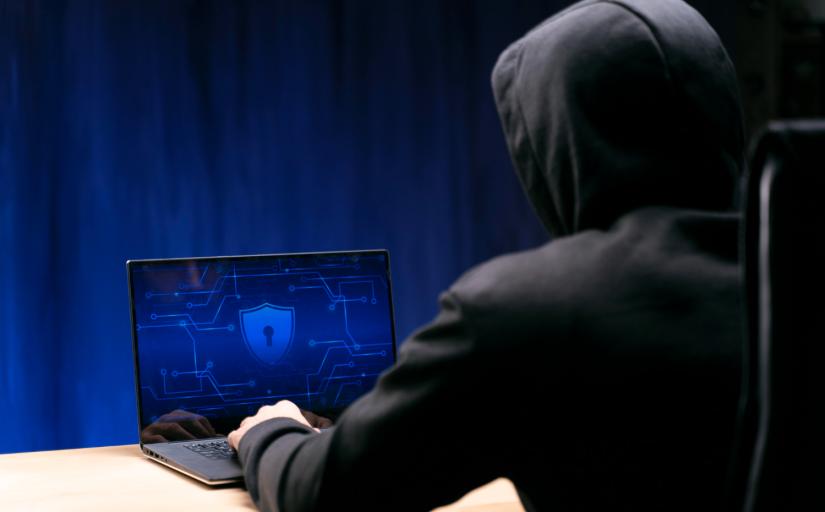 Foto de um homem vestido com um moletom preto com capuz, de costas, sentado. Na frente um laptop com uma imagem no tom azul