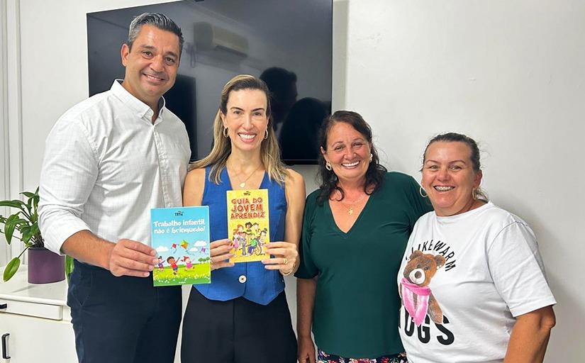 Qautro pessoas, uma ao lado da outra. à esquerda, um homem alto, de camisa branca, pele clara e cabelos grisalhos, em seguida uma mulher loira, de blusa azul,  ao lado uma mulher morena, de cabelos longos e blusa verde e em seguida uma mulher morena, de cabelos presos, de camiseta branca