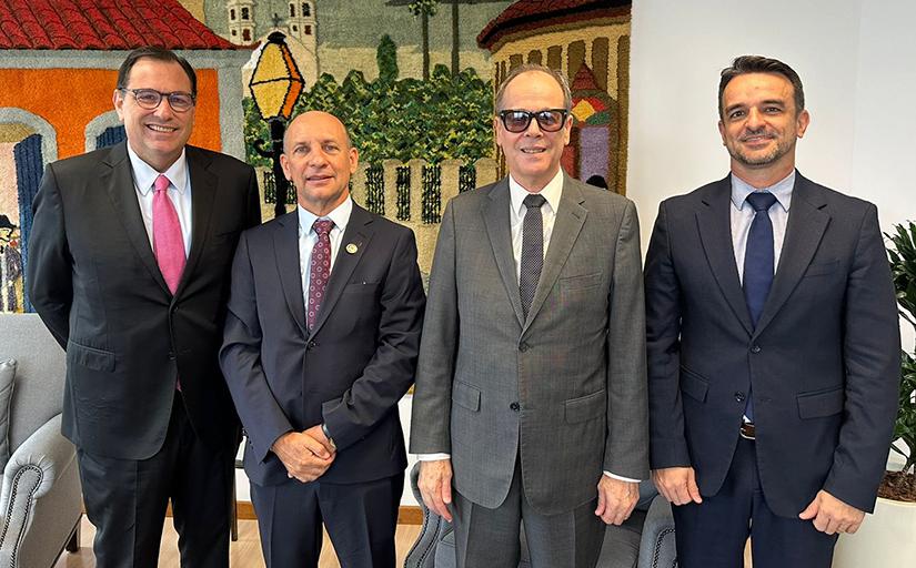 Quatro homens de terno escuro posam de pé para a foto, sorridentes. Ao fundo, um quadro colorido.