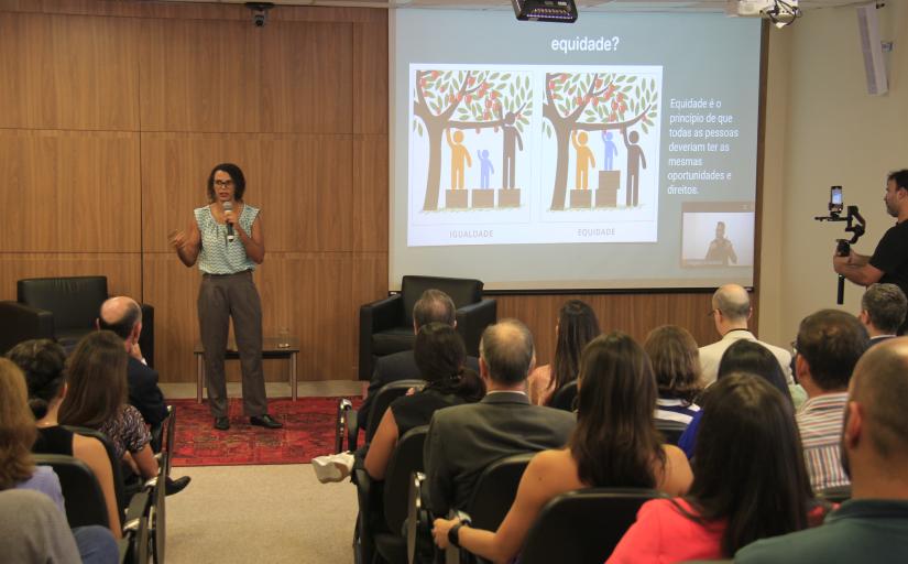 Foto de uma palestra com uma mulher negra ao lado de um telão, falando ao microfone, e pessoas sentadas no auditório