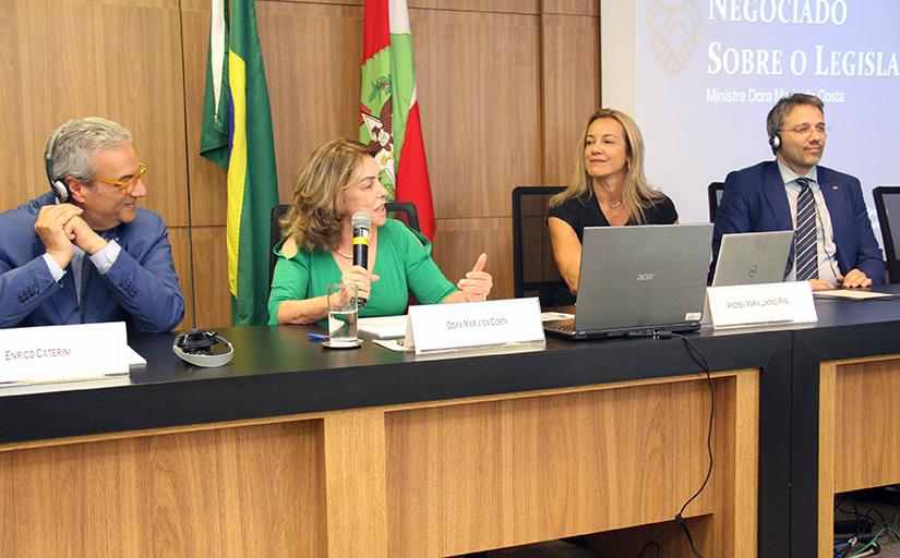 Quatro pessoas, sendo dois homens e duas mulheres, estão sentados em uma mesa no auditório do tribunal, com a bandeira do Brasil, de Santa Catarina e uma tela de projeção ao fundo