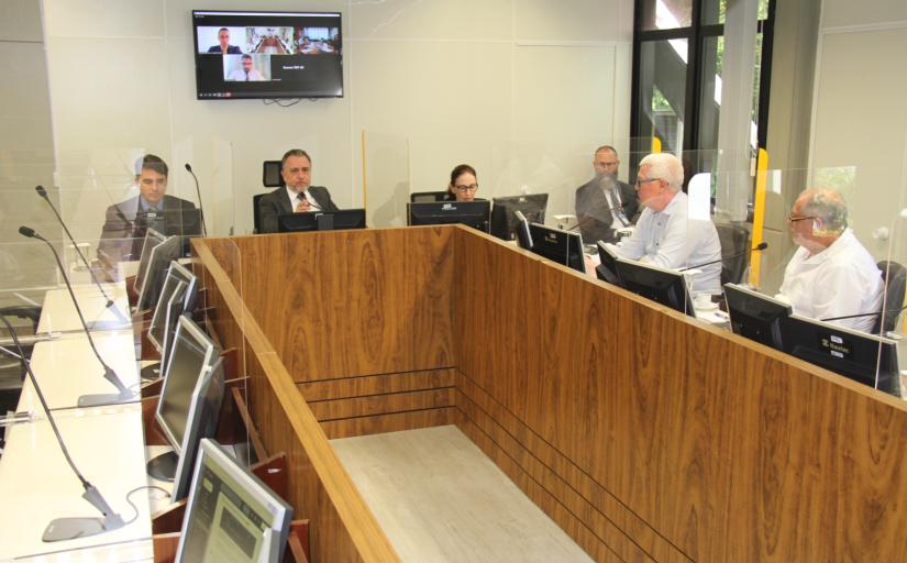 Fotografia da audiência na Seção Especializada 1 (SE-1) do TRT-SC. Em volta de uma bancada de madeira, quatro homens e uma mulher estão vestidos formalmente, diante de monitores de computador. Ao fundo, um monitor exibe rostos.