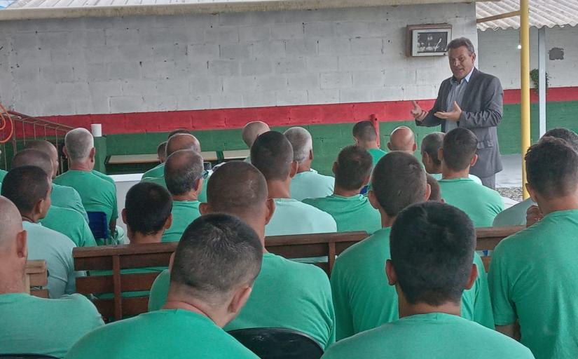 Homem ao fundo de terno cinza, e à sua frente diversos homens de camiseta verde de costas para a foto