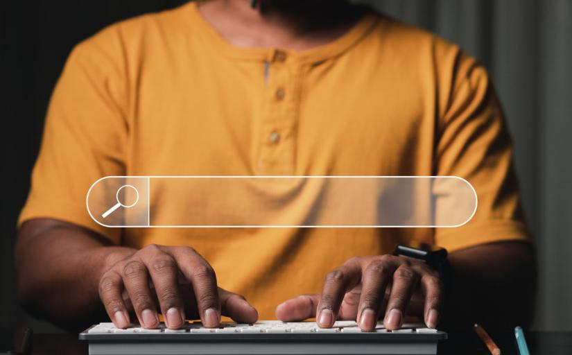 Parte de um homem negro aparece em uma foto. Ele veste camisa laranja e digita num teclado de computador