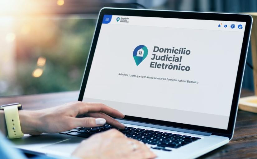 Foto de banco de imagens. Mãos de uma pessoa manuseando um notebook sobre uma mesa, fundo desfocado. Na tela do computador há o logo do Domicílio Judicial Eletrônico.