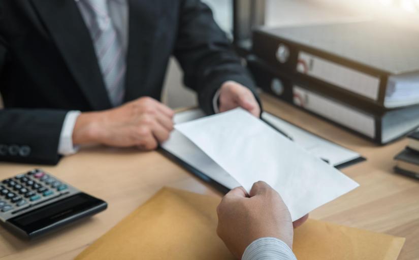 foto ilustrativa de uma mão dando um envelope a um homem de terno sentado em uma mesa de escritório