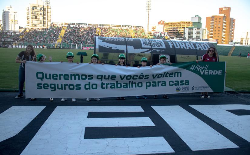 crianças com capacetes verdes segurando uma faixa com os dizeres "queremos que vocês voltem seguros para casa"