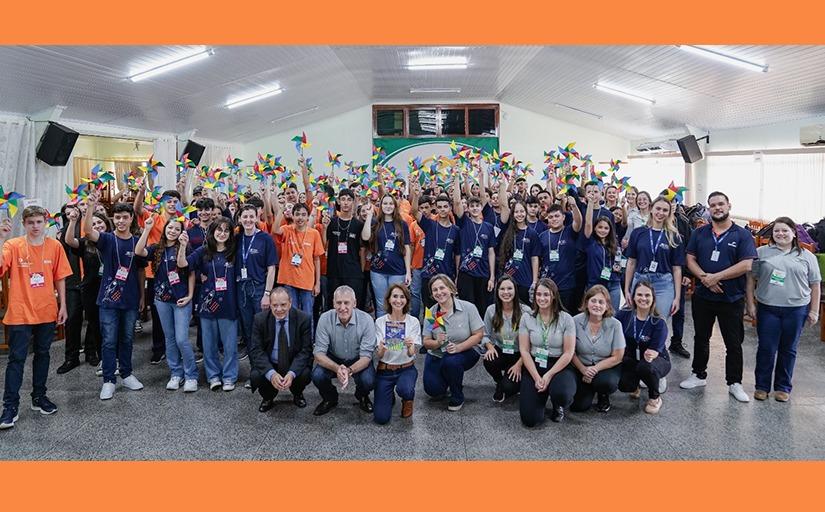 Grupo de cerca de 100 jovens, vestindo uniformes nas cores azul e laranja, posam para foto com braços levantados e segurando cataventos, em uma sala ampla