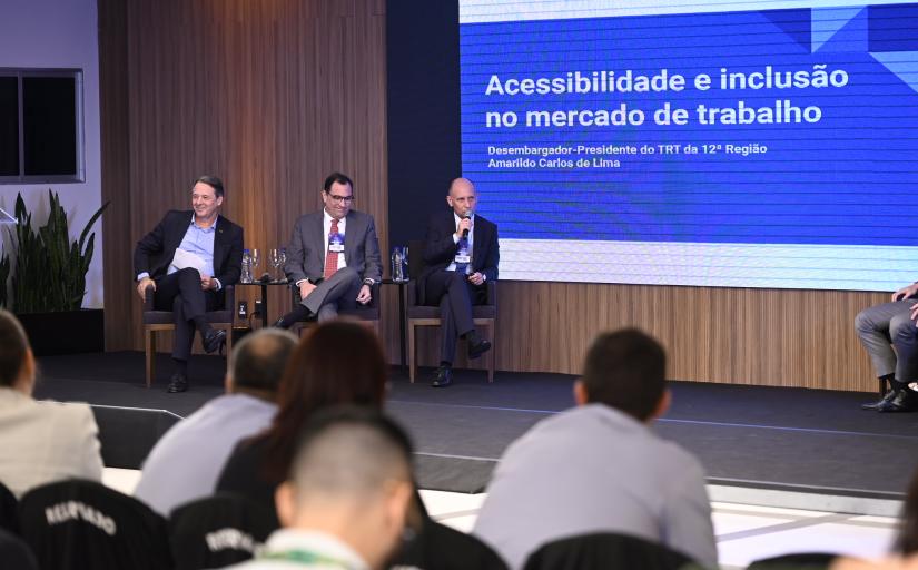 Três homens brancos, de cabelos escuros, trajando ternos, estão sentados em cadeiras em um palco. Um deles, à direita, é o desembargador Amarildo Carlos de Lima, que fala ao microfone. Em uma tela ao fundo se lê: Acessibilidade e inclusão no mercado de trabalho. Desembargador-presidente do TRT da 12ª Região, Amarildo Carlos de Lima.