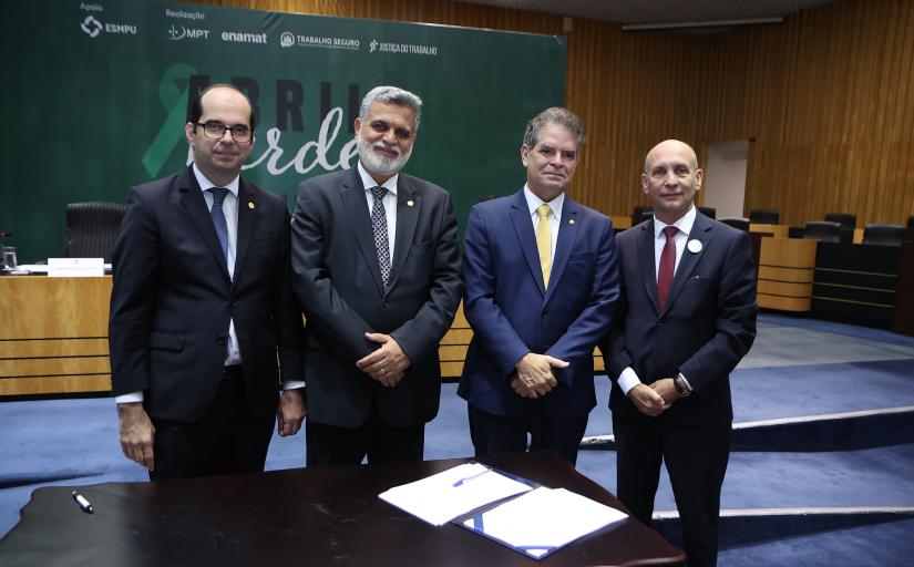 Foto mostra quatro homens, todos de terno escuro, de pé e sorridentes. Eles estão em um auditório e, à frente deles, está uma mesa com alguns papéis e uma caneta sobre ela.