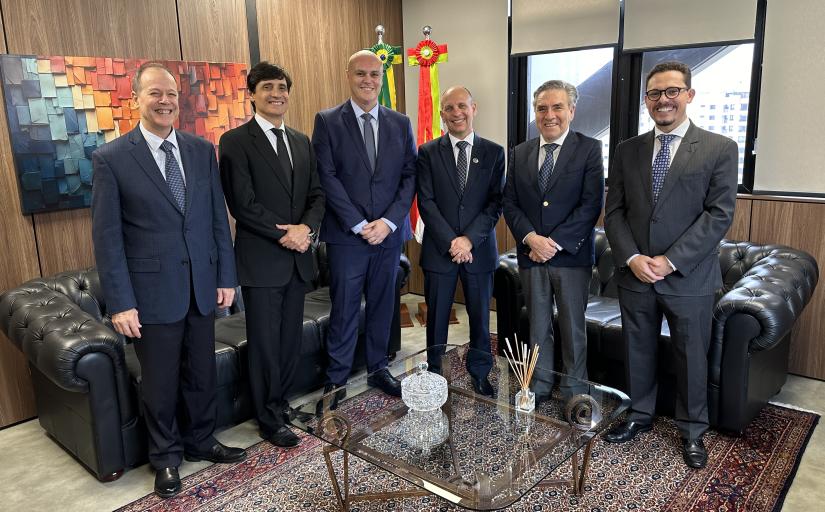 Seis homens em pé e de terno e gravata, posando para a foto, dentro de uma sala.