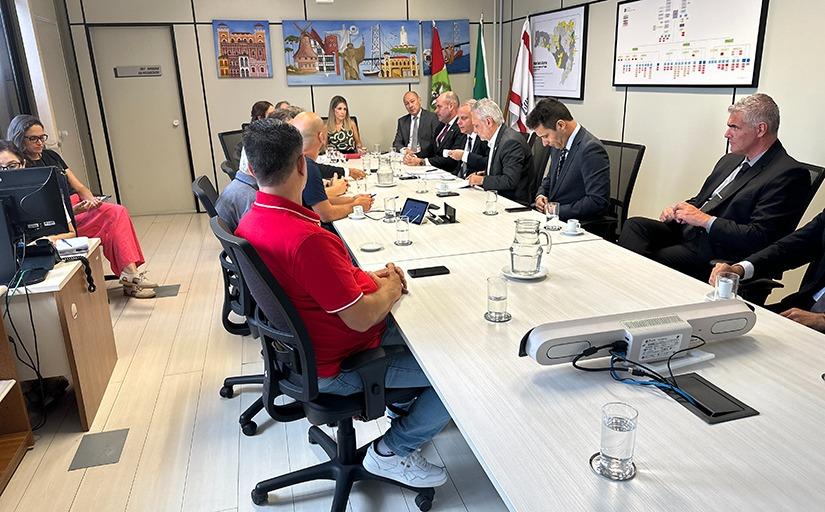 Foto mostra 14 pessoas em torno de uma mesa horizontal, branca, e longa. À direita, seis homens de terno; à esquerda, sete homens vestidos informalmente e uma mulher, mais forma. Na cabeceira, uma mulher loira observa a conversação.