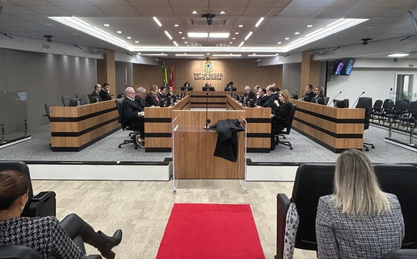 fotografia da sala de sessões do tribunal pleno do TRT-SC. várias pessoas de toda reunidas em torno de bancadas de madeira. é possível ver também uma parte do auditório, com pessoas sentadas