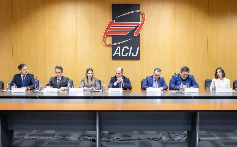 Foto mostra cinco homens e duas mulheres sentados em uma mesa de evento. Ao fundo, o logo da Associação Comercial e Industrial de Joinville