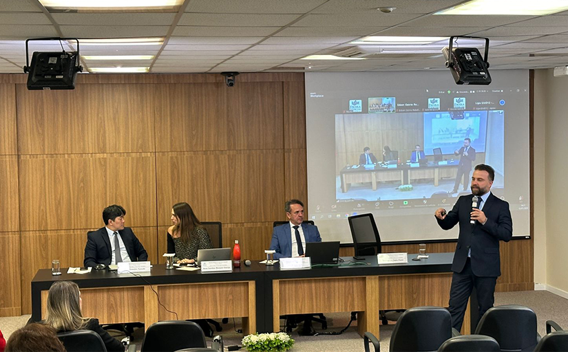 Foto de uma apresentação em um auditório. Um homem branco de cabelo curto, barba e bigode escuro, vestindo terno preto, fala levando um microfone à boca. Atrás dele está uma mesa onde se encontram uma mulher e dois homens sentados e um projetor que transmite uma reunião virtual com enquadramento similar ao da foto.