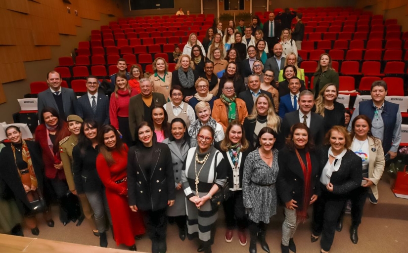 Foto mostra um grupo de cerca de 60 pessoas de pé, agrupadas, a maioria composta por mulheres, posando sorridente para a câmera. A foto é registrada de cima para baixo e elas estão em um auditório com assentos vermelhos
