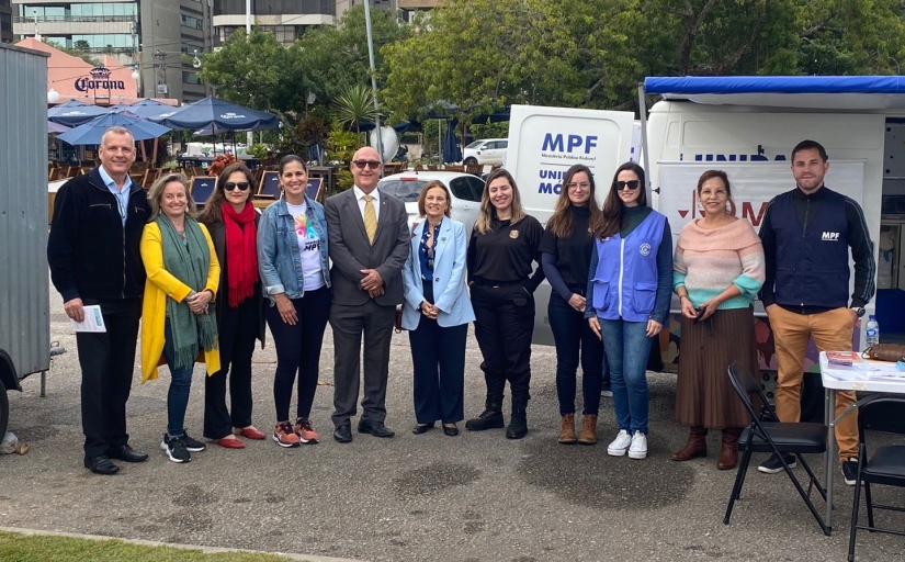 Foto de onze pessoas em um ambiente externo. Atrás de veem carros estacionados, um quiosque e uma van com as portas traseiras abertas, na porta está escrito "MPF"