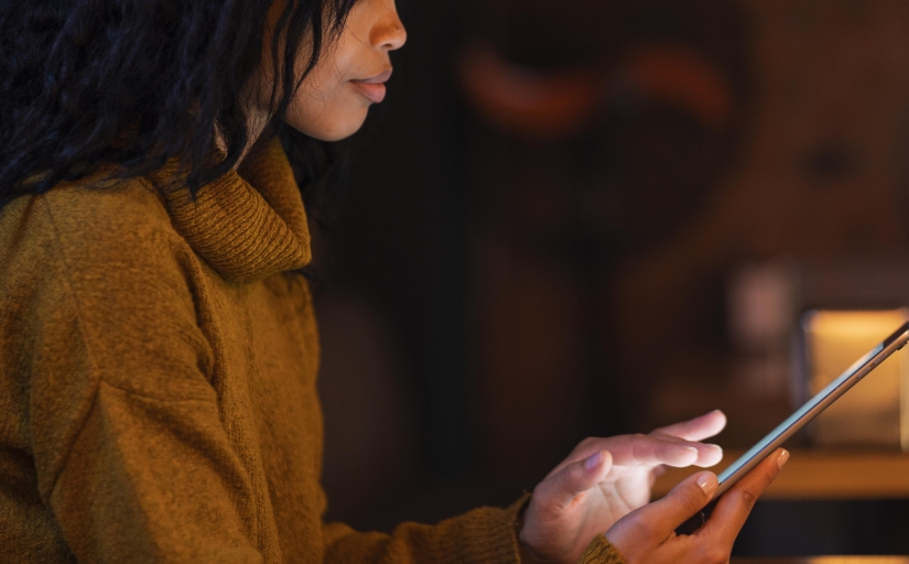 Foto de uma mulher negra, de perfil, vestindo um blusão marrom claro, usando um tablet que está iluminado