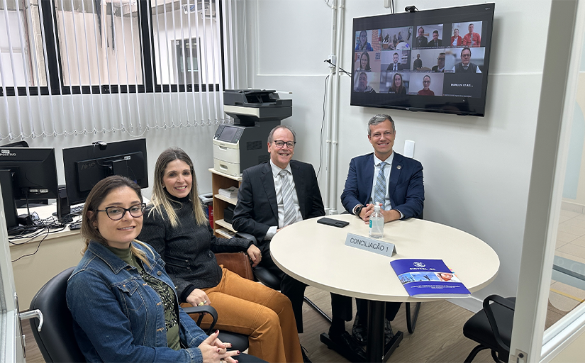 Foto de duas mulheres e dois homens sentados em uma mesa redonda, posando para a foto. Ao fundo uma tela de TV com uma reunião virtual