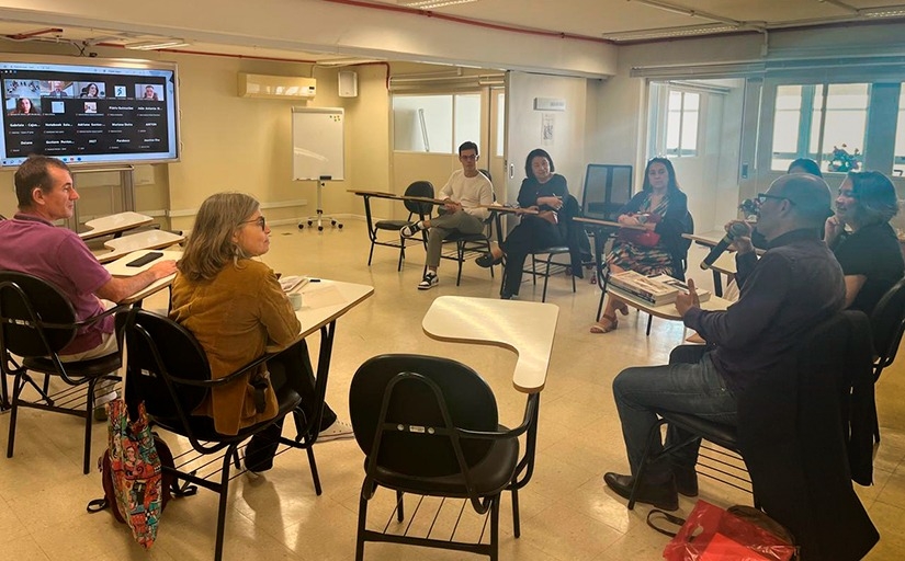 Fotografia do evento. Várias pessoas estão sentadas reunidas em uma roda, enquanto um deles segura o microfone e fala. Ao fundo, um telão mostra participantes online.