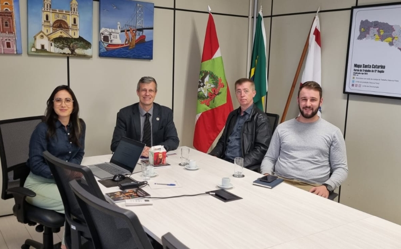 Quatro pessoas sentadas ao redor de uma mesa de reunião. À esquerda, uma mulher morena, de óculos, usando um terno azul escuro, ao lado um homem grisalho, de terno preto, em seguida um homem branco, de olhos azuis e cabelo ruivo, vestindo uma camisa azul e uma jaqueta preta e ao seu lado, um homem branco, de barba e cabelos castanhos, vestindo uma blusa cinza