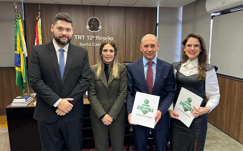 Duas mulheres e dois homens em pé, posando para a foto, em um gabinete. Ao fundo, bandeiras do Brasil e o brasão da Justiça do Trabalho plotado em um MDF marrom escuro