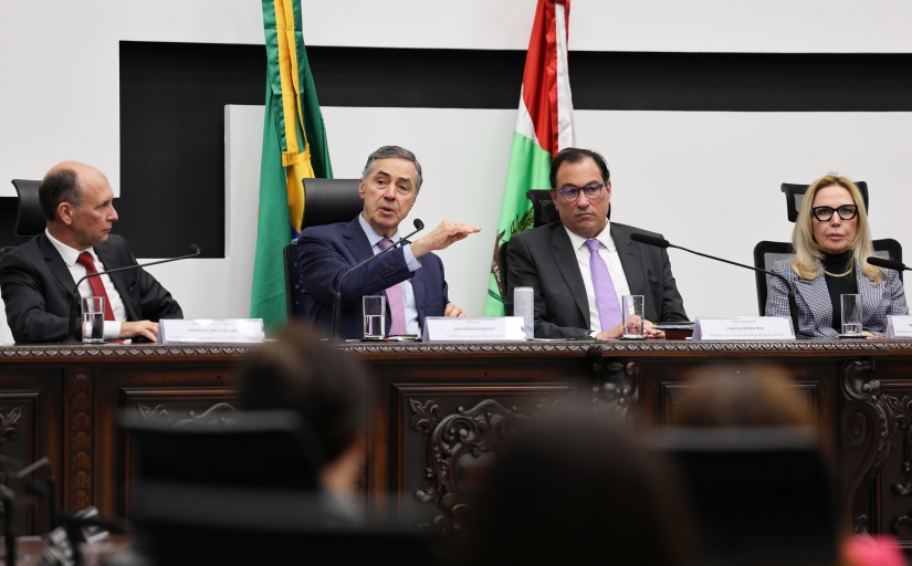 Três homens e uma mulher estão sentados em uma mesa, todos trajando roupas formais. Um deles, o ministro do STF Luís Roberto Barroso, fala ao microfone.