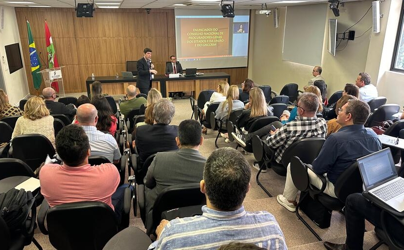 Fotografia de um auditório cheio. À frente, falando para os presentes, um homem de terno segura o microfone. Ao fundo, sentado atrás de uma bancada de madeira, há outro homem de terno