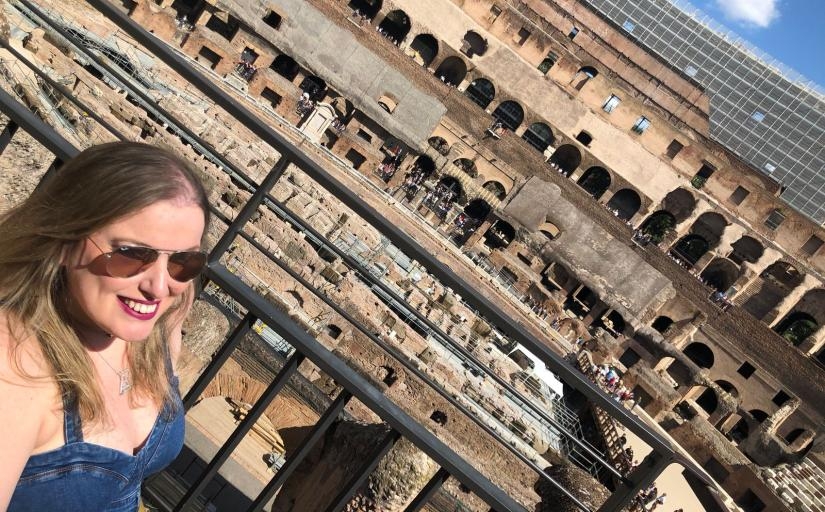 Uma mulher loira com óculos escuros sorri para a foto. Ao fundo o Coliseu, em Roma