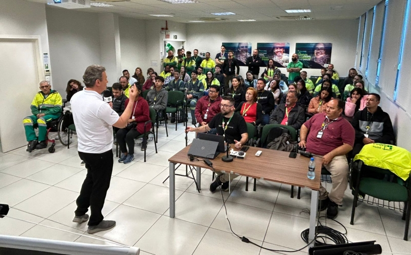 A imagem mostra uma palestra em um ambiente interno, onde um palestrante, de camisa branca, está falando para uma plateia de pessoas sentadas em cadeiras.A audiência é composta por indivíduos vestidos com uniformes verdes e amarelos, Alguns estão de pé ao fundo. Há equipamento de projeção no teto e uma mesa com computadores e materiais à frente. 