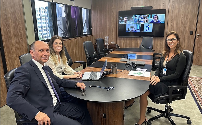 A imagem mostra três pessoas sentadas ao redor de uma mesa de conferência oval em uma sala bem iluminada. Na tela grande ao fundo, aparecem quatro pessoas participando de uma videoconferência. Na mesa, há um laptop aberto, papéis, um telefone e um par de óculos. O ambiente sugere um encontro profissional, possivelmente uma reunião de negócios ou organizacional, com participantes presenciais e virtuais.